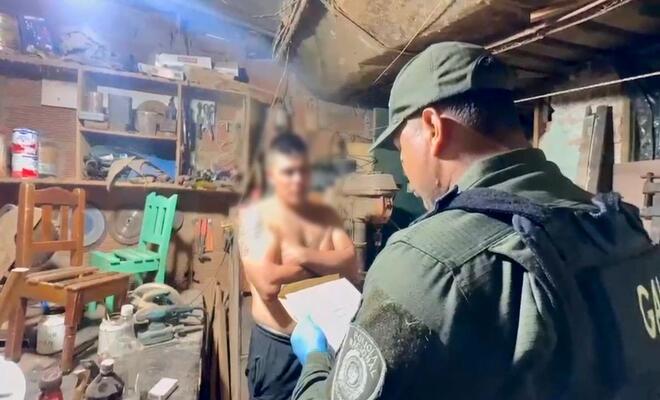 Interior de un taller con herramientas y estanterías; una persona uniformada observa un documento mientras otra persona se encuentra de pie frente a ella, y objetos de trabajo están distribuidos sobre mesas y paredes.