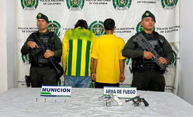 Dos hombres capturados en medio de dos Polic&iacute;as, en frente armas de fuego incautadas.