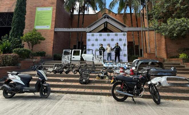 Escalinata exterior frente a un edificio institucional con un fondo de la Policía Nacional de Colombia; en primer plano hay motocicletas, una camioneta y múltiples autopartes organizadas sobre los escalones, mientras varias personas se encuentran de pie detrás.