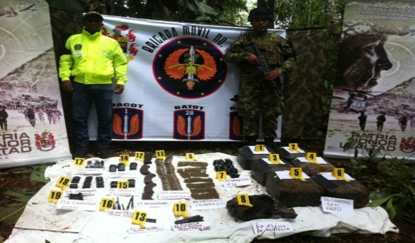 fotografía con un policía y un soldado parados, y adelante de ellos material de guerra incautado