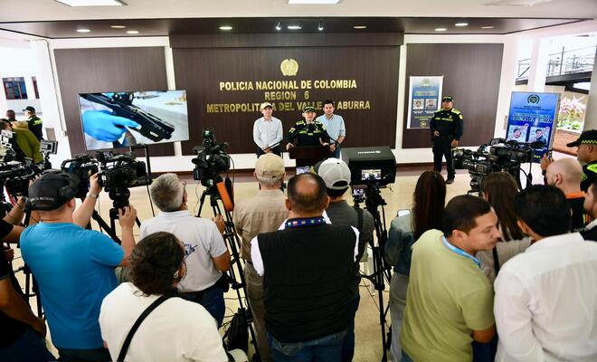 Sala de prensa con varias personas de pie frente a un fondo institucional; en primer plano se observan cámaras y equipos de grabación dirigidos hacia una mesa principal donde se realiza una intervención vocero de la Policía Nacional.